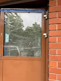 Brązowe drzwi aluminiowe z osadzoną szybą zabezpieczoną taśmą, przy ceglanej elewacji.