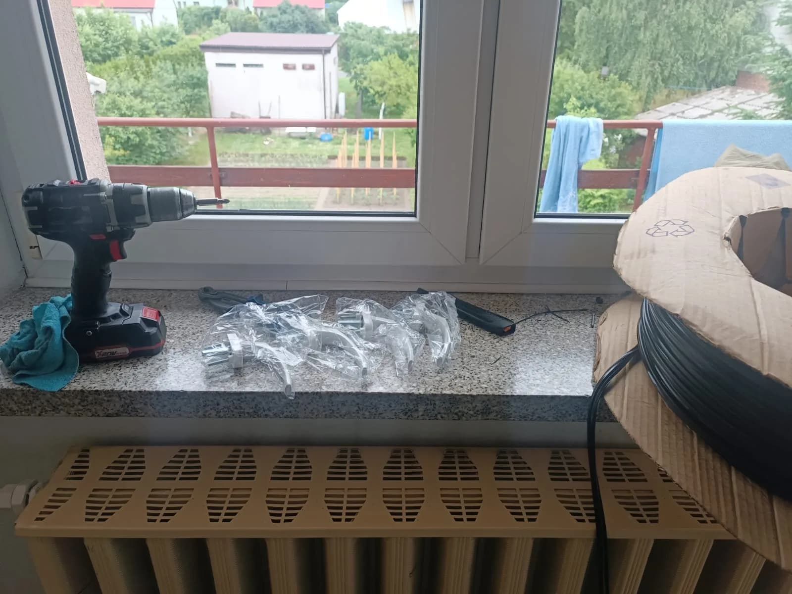 Nowe okucia okienne w opakowaniu i wkrętarka na parapecie przy oknie.