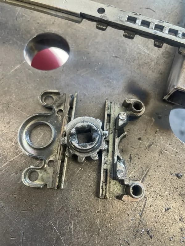Rozłożony mechanizm zasuwnicy okiennej z gniazdem klamki na blacie warsztatowym.