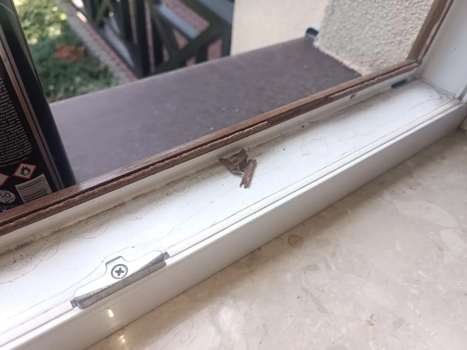 Sparciała brązowa uszczelka i odłamane fragmenty przy metalowym zaczepie okucia okna.