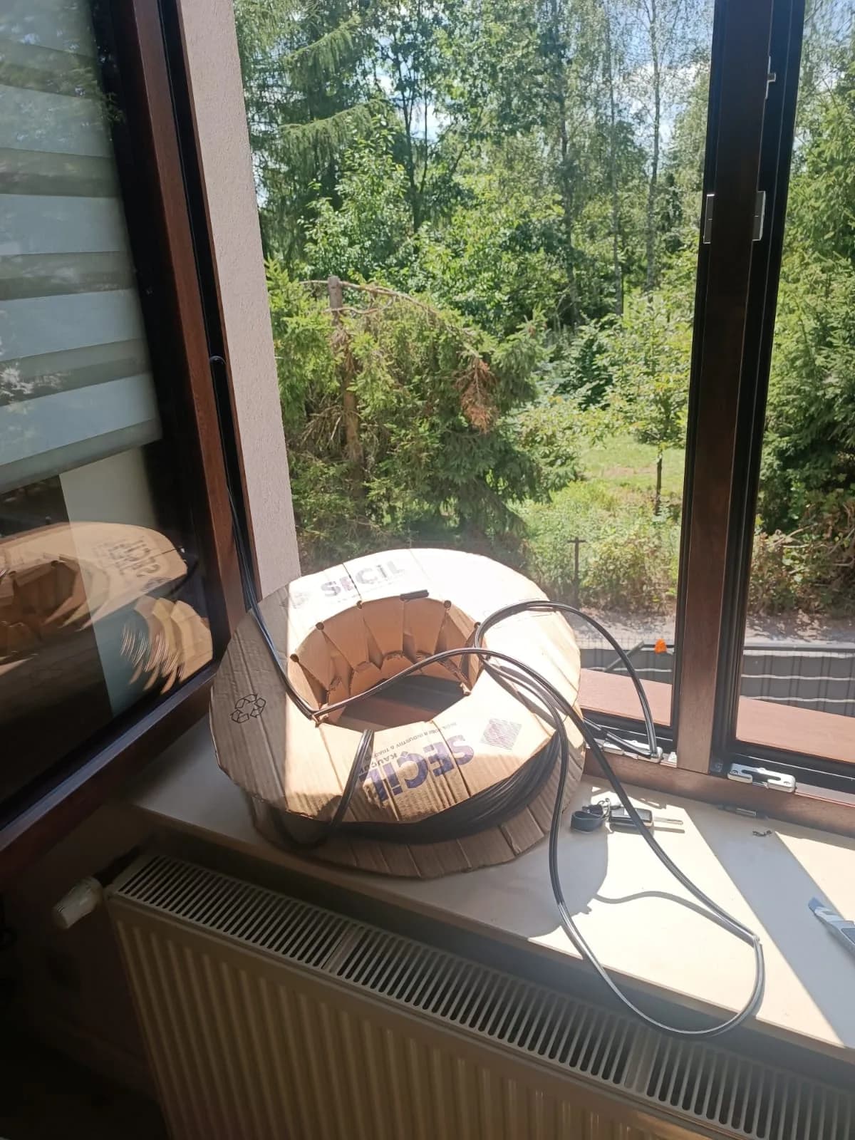 Szpula czarnej uszczelki do okna drewnianego ustawiona na parapecie przy otwartym skrzydle.
