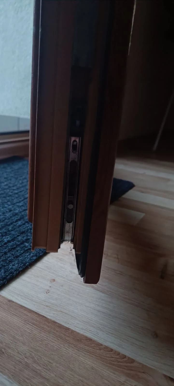 Uszkodzony dolny narożnik brązowych drzwi balkonowych z odłamanym fragmentem profilu.