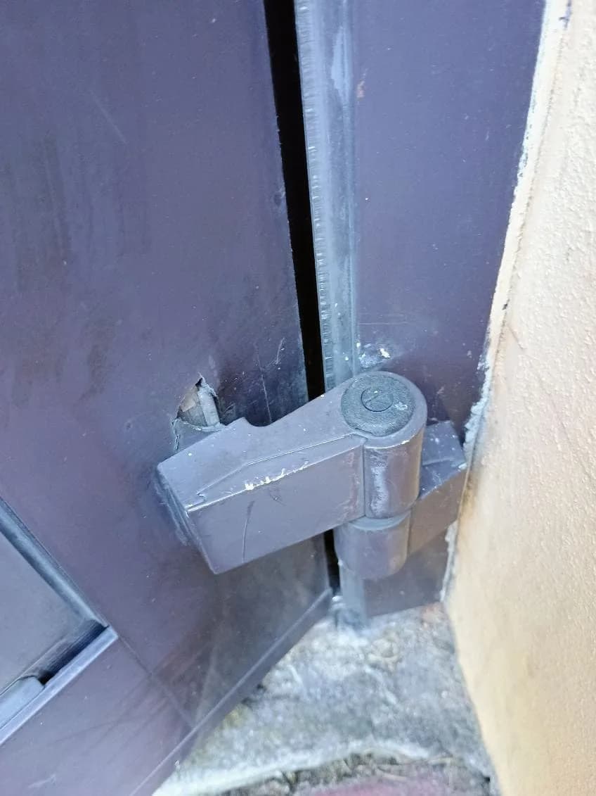 Uszkodzona obudowa zawiasu w brązowych drzwiach aluminiowych z widocznym pęknięciem.